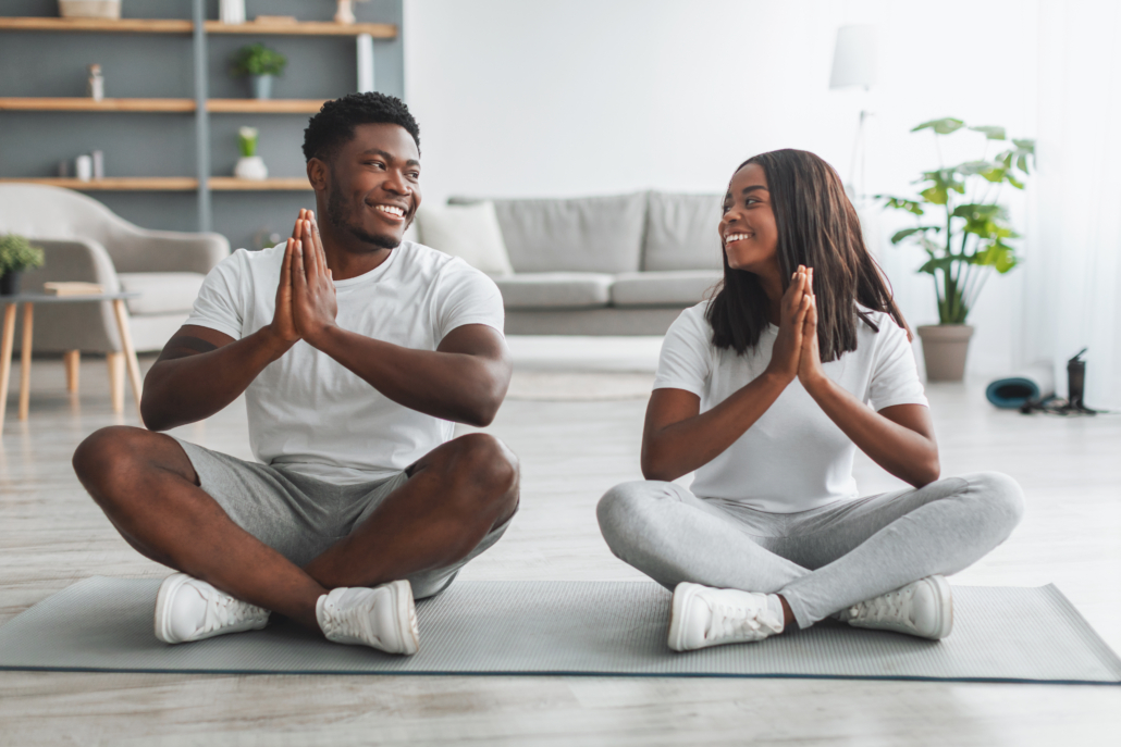 Portrait of happy smiling young black couple holding hands in pray pose, keeping palms together, breathing and meditating, sitting on floor mat.
